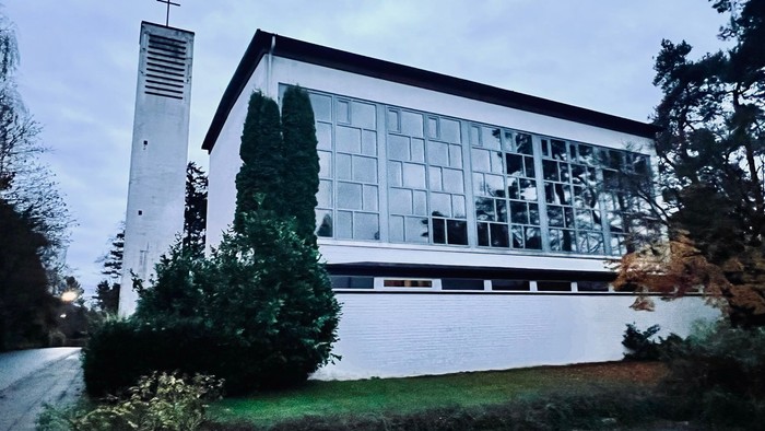 Moderne Kirchengebäude mit großen Glasfenstern und einem hohen Glockenturm neben üppigem Grün.