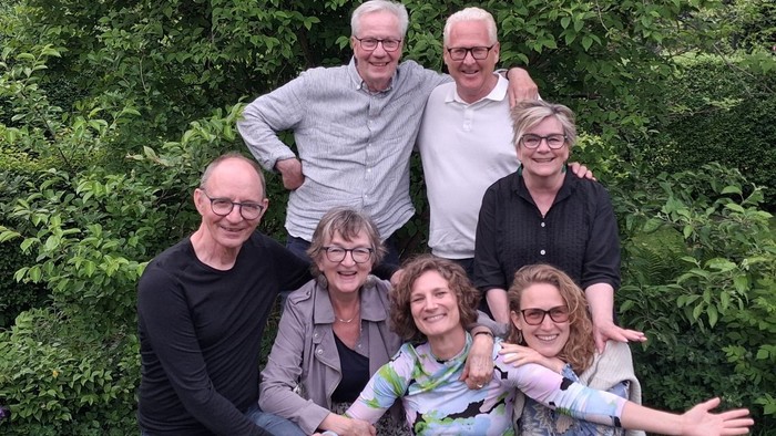 En glad gruppe på syv personer, der poser sammen udendørs blandt grønt løv.