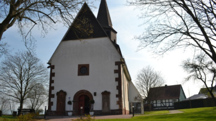 Idyllische weiße Kirche mit steilem Satteldach und dunklem Turm, umgeben von kahlen Winterbäumen und einer grasbewachsenen Fläche.