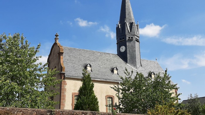 Kirche mit hohem Turm und Uhr, umgeben von Bäumen und Blumen.