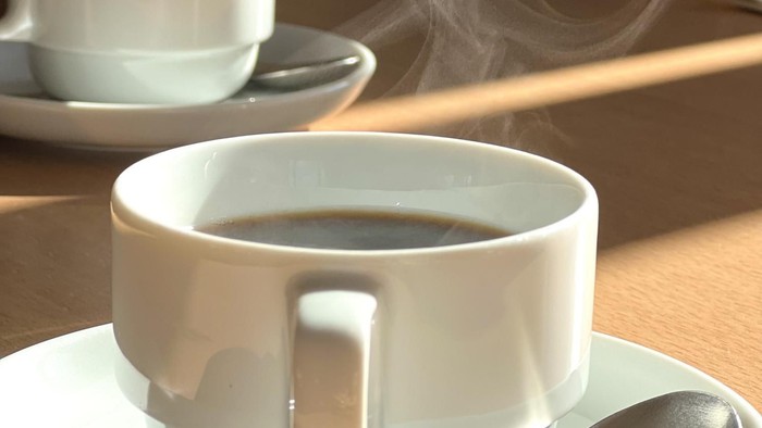 Heißer dampfender Kaffee in einer weißen Tasse mit einem Löffel auf einer Untertasse, platziert auf einem Holztisch.