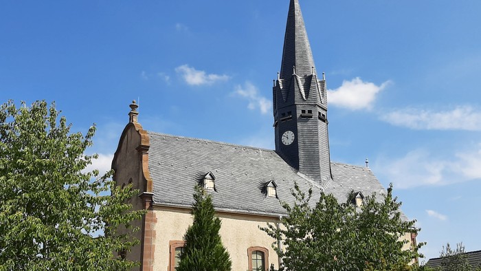Kirche mit hohem Turm und Uhr, umgeben von Bäumen und Blumen.