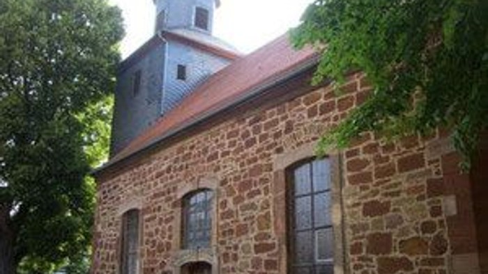 Historische Steinkirche mit Glockenturm und Bogenfenstern, umgeben von Grün.