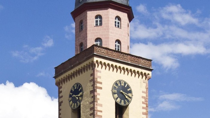 Hoher Kirchturm mit Uhr und Wetterfahne, davor Café mit Sonnenschirmen