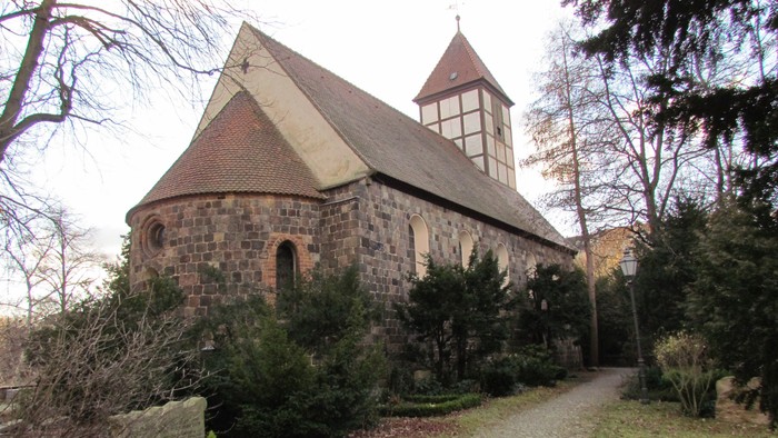 Kleine Kirche mit Turm und steilem Dach, umgeben von Bäumen und Büschen