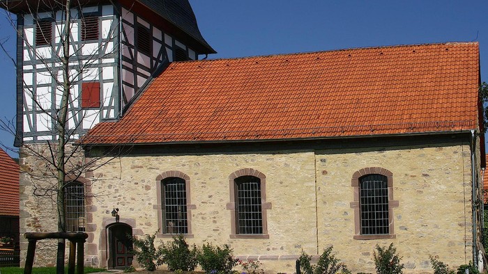 Außenansicht der evangelischen Kirche in Vernawahlshausen. Natursteinkirchenschiff mit rotem Ziegeldach und Fachwerkturm.