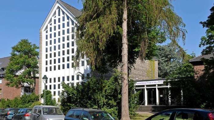 Städtische Szene mit einem hohen weißen Gebäude, vor dem geparkte Autos und Grünflächen vor einem klassizistischen Eingang zu sehen sind.