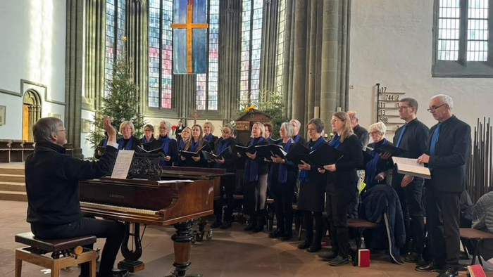 Ein Chor probt in einer prächtigen Kirche, angeführt von einem Pianisten, mit bunten Kirchenfenstern im Hintergrund.
