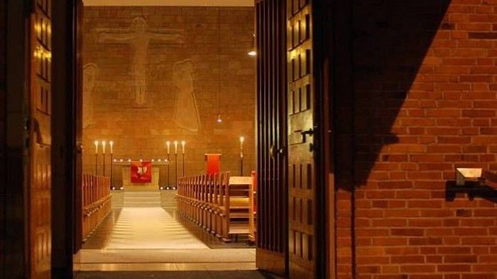 Warm beleuchteter Kirchengang mit Bänken und religiösen Kunstwerken an der Wand, eingerahmt von Backsteinmauern und Türen.