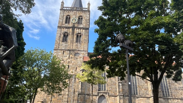 Das Bild zeigt links den großen Glockenturm aus Sandstein und nach rechts das weitere Kirchengebäude mit hohe Fenstern, teilweise von Bäumen im Vordergrund verdeckt.
