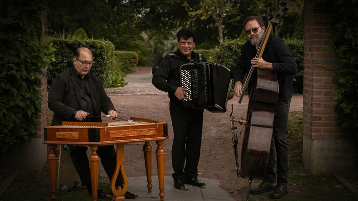 Tre musikere, der spiller på harmonika, keyboardharpe og kontrabas udendørs i en have.