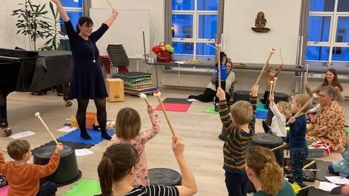 En gruppe børn og en instruktør deltager i et musikalsk aktivitet i en lys og åpen lokalitet.