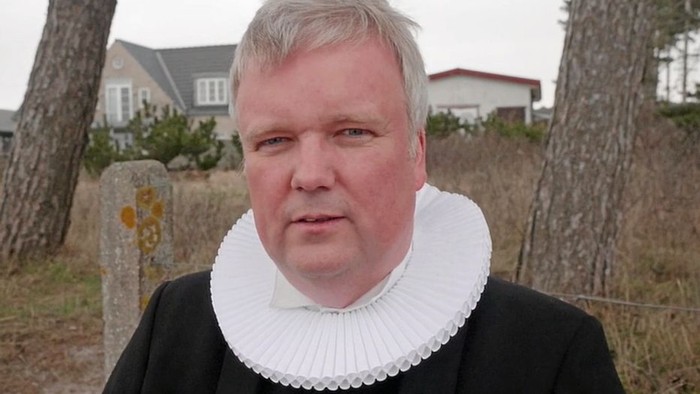 En mand i formelt tøj med en krusekrave står udendørs nær et boligområde med træer.