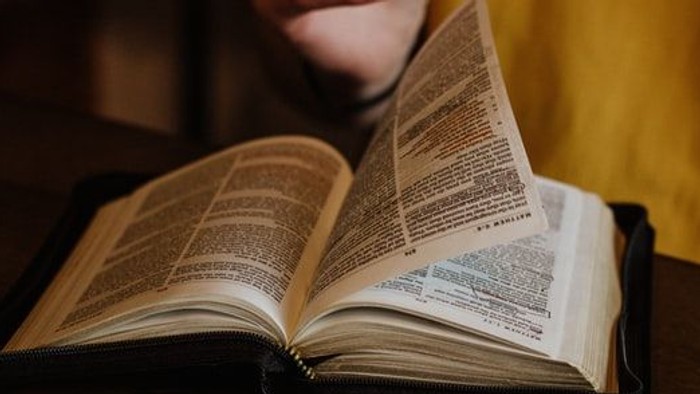 Eine Person liest ein aufgeschlagenes Buch mit gefalteten Händen über den Seiten auf einem Holztisch.