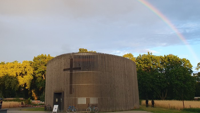 Ein beeindruckender Regenbogen wölbt sich über ein rustikales Holzgebäude, umgeben von üppigem Grün und abgestellten Fahrrädern.
