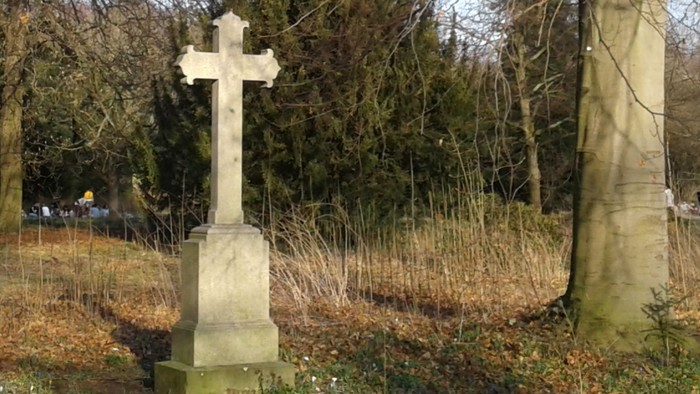 Ein Grabkreuz steht in einem bewaldeten Bereich mit Bäumen und trockenem Laub.