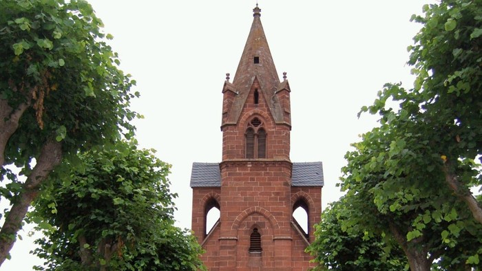 Kirche mit hohem Turmspitz und Rundbogenfenstern, umgeben von Bäumen.