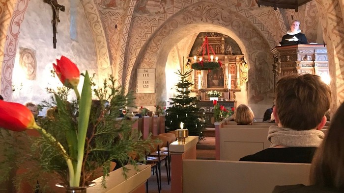 Et roligt kirkeinteriør med kirkebænke, festlige dekorationer og religiøs kunst på væggene under en gudstjeneste.