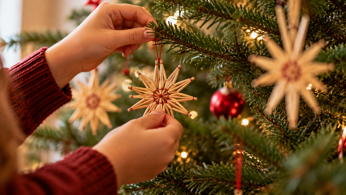 Zwei Hände hängen einen Strohstern an einen mit Lichterkette, roten Kugeln und weiteren Strohsternen geschmückten Weihnachtsbaum.