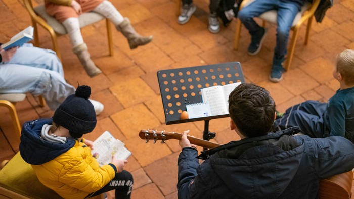Eine Gruppe versammelt sich in einem gemütlichen Raum, singt und spielt Gitarre mit Notenblättern auf einem Notenständer.
