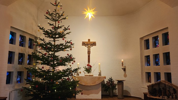 Altarraum mit geschmücktem Tannenbaum, Krippe und brennenden Kerzen. Oben leuchtet ein Stern.