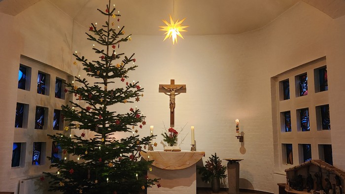 Altarraum mit geschmücktem Tannenbaum, Krippe und brennenden Kerzen. Oben leuchtet ein Stern.