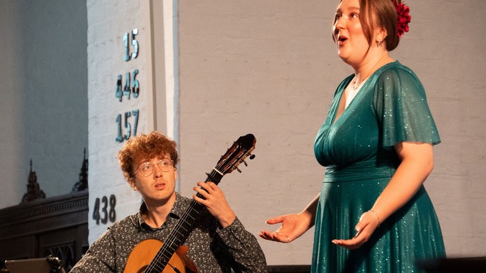 To optrædende synger og spiller guitar på scenen med et nummereret bagtæppe.