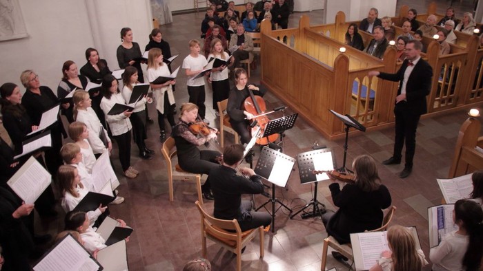 Ein Chor und ein kleines Orchester treten gemeinsam in einer Kirche auf, dirigiert von einem Dirigenten von vorne.