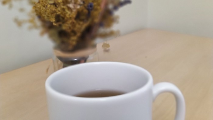 Eine weiße Tasse Tee auf einem Holztisch mit einer bunten Deko und getrockneten Blumen im Hintergrund.