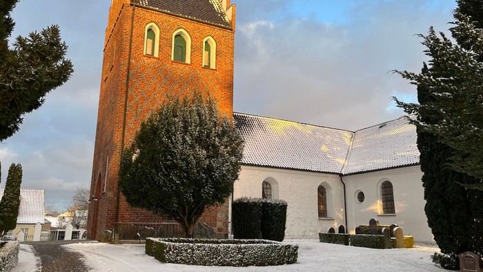 En sneklædt kirke med et højt murstenstårn og hvide mure, omgivet af stedsegrønne træer og en klar himmel.