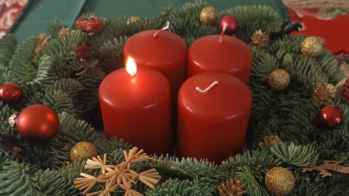 Auf einer grünen Tischdecke steht ein Adventskranz mit roten Kerzen. Die erste Kerze brennt. 