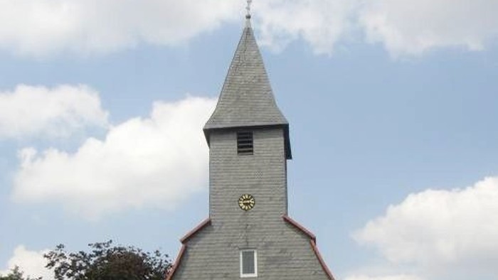 Kleine Steinkirche mit hohem Turm und Uhr, umgeben von Grünflächen.