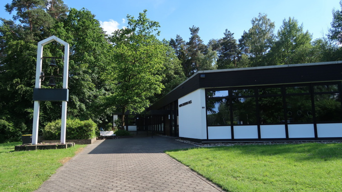 Modernes Gebäude mit großen Fenstern, umgeben von üppigem Grün und einer hohen weißen Struktur neben einem gepflasterten Weg.