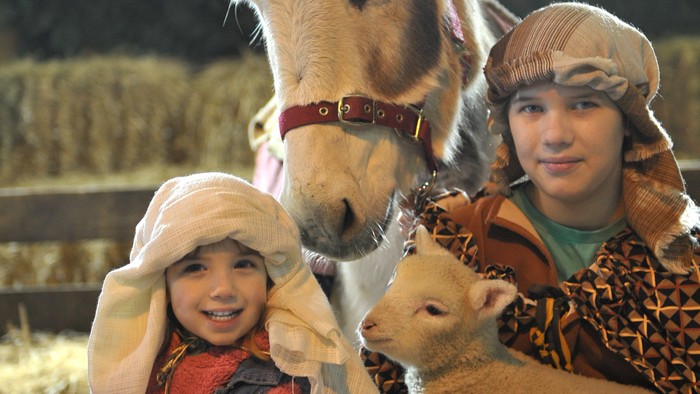 Children pose with donkey and lamb, dressed in rustic clothing, under string lights.