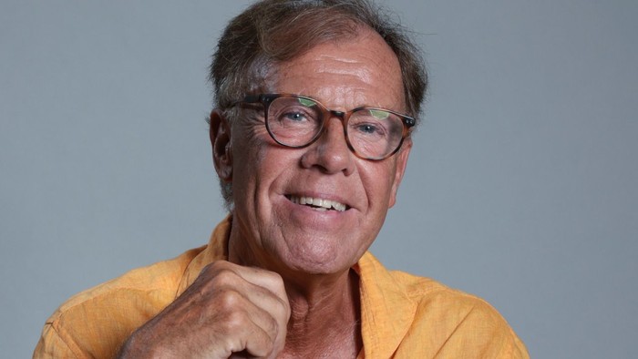 Mand med et smil i en gul skjorte poser selvsikkert med hånden nær hagen.