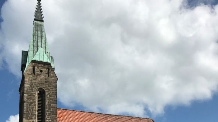 Historische Steinkirche mit steilem rotem Ziegeldach und hohem Turm unter einem teilweise bewölkten Himmel.