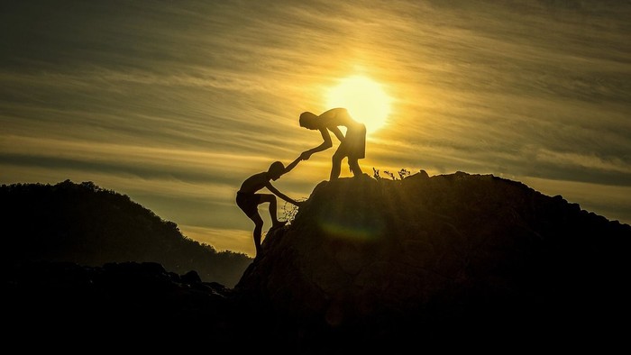 Silhouetten von zwei Personen, die bei Sonnenuntergang einen felsigen Hügel erklimmen.
