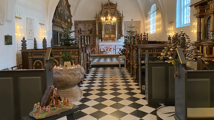 Kirkeinteriør med sort-hvid skakbrætgulv, religiøse dekorationer og krystallysekroner.
