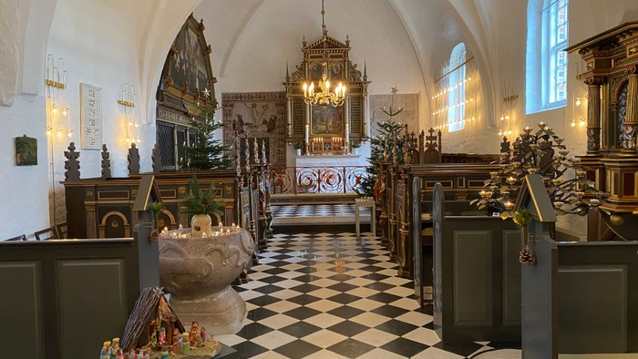 Kirkeinteriør med sort-hvid skakbrætgulv, religiøse dekorationer og krystallysekroner.