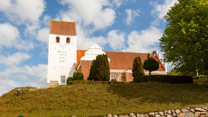 En hvid kirke med røde tag og tårn på en bakke med træer og stenmur.