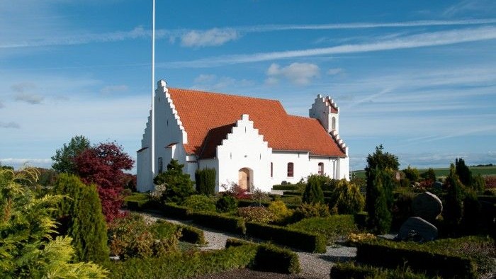 En hvid kirke med rødt tag og en flagstang foran, omgivet af grønne planter og buske