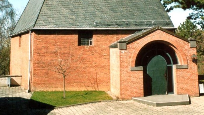 Idyllische Backsteinkapelle mit steilem dreieckigem Dach und markantem Kreuz an der Spitze.