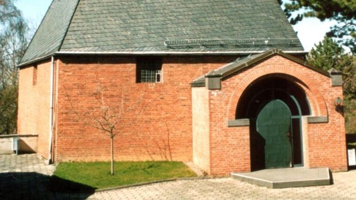 Idyllische Backsteinkapelle mit steilem dreieckigem Dach und markantem Kreuz an der Spitze.