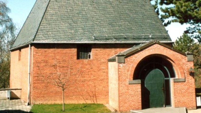 Idyllische Backsteinkapelle mit steilem dreieckigem Dach und markantem Kreuz an der Spitze.