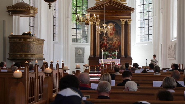 In der Kirche sitzen Menschen in Holzbänken, vorne Altar mit Kerzen und Gemälde.