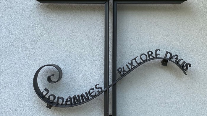 Das Bild zeigt ein schwarzes Kreuz mit lateinischem Text an einer weißen Wand.