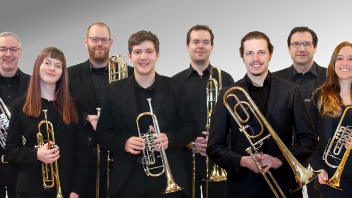 Musikerinnen und Musiker eines Blechbläserensembles stehen nebeneinander vor hellem Hintergrund. Sie tragen schwarze Konzertkleidung und halten verschiedene Blechblasinstrumente wie Trompeten und Posaunen in den Händen. Alle blicken in die Kamera und läch