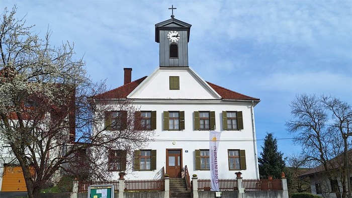Weißes Gebäude mit Uhrturm und Kreuz, umgeben von Bäumen