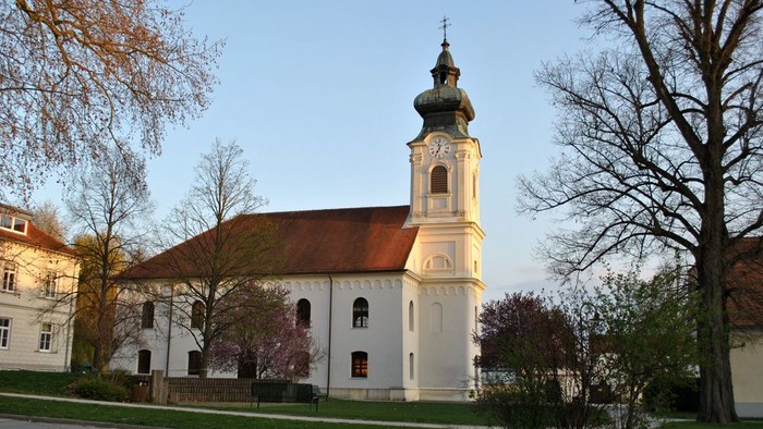 Weiße Kirche mit hohem Turm und roter Dachziegeln, umgeben von Bäumen und einem klaren blauen Himmel