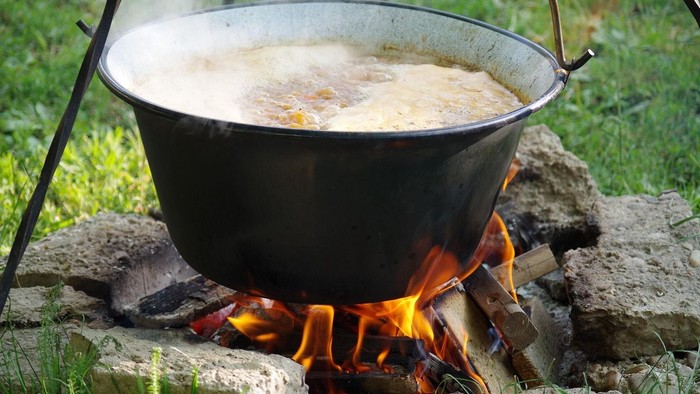 Ein großer Topf köchelt über einem offenen Lagerfeuer, während Dampf aus seinem Inhalt aufsteigt.